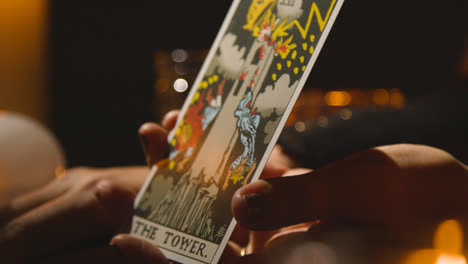 Close-Up-Of-Woman-Giving-Tarot-Card-Reading-On-Candlelit-Table-Holding-The-Tower-Card-3